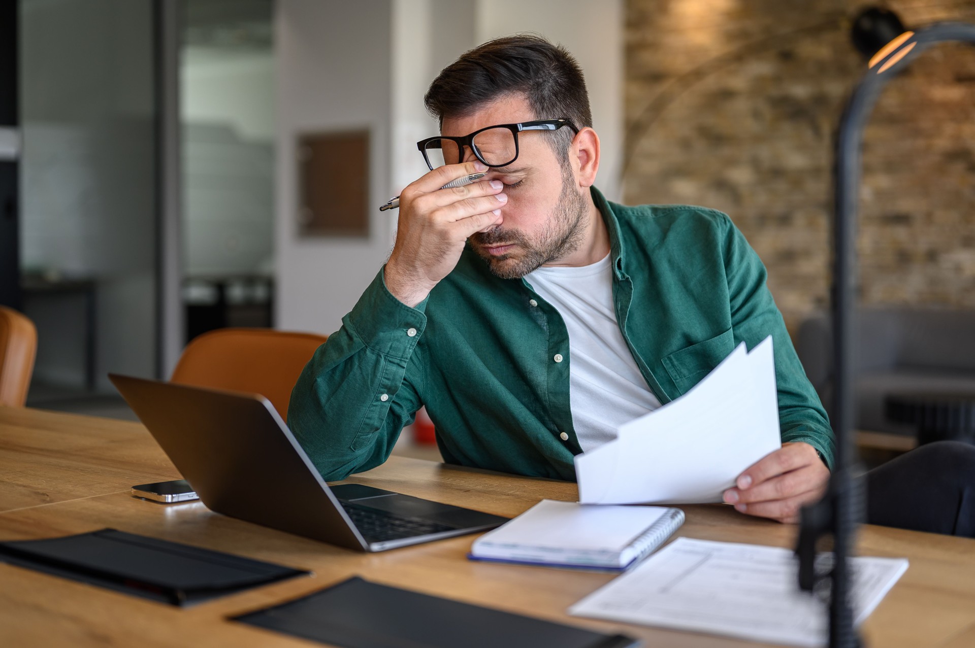 Consulente finanziario maschio ansioso che si strofina gli occhi mentre lavora sul laptop e analizza i rapporti alla scrivania